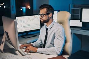 Selbstbewusster junger Mann, der am Computer arbeitet, während er lange im Büro bleibt foto