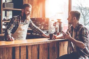 gutes gespräch mit kunden. barista und sein kunde diskutieren etwas mit einem lächeln, während sie an der bartheke im café sitzen foto