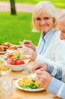 Essen mit dem nächsten. Älteres Paar, das beim Sitzen am Esstisch im Freien isst und lächelt foto