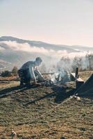 wichtiger Bestandteil des Campings. Junger Mann, der ein Lagerfeuer macht, während er in der Nähe des Zeltes in den Bergen sitzt foto