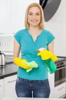 Hausfrau bei der Arbeit. Reife Frau mit blonden Haaren in gelben Handschuhen, die einen Lappen und eine Sprühflasche hält, während sie in die Kamera lächelt foto