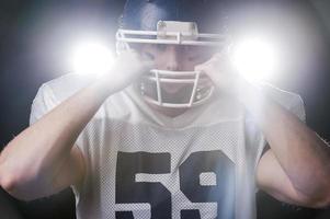 ernsthaftes Spiel beginnt. Porträt eines American-Football-Spielers, der die Hände auf dem Helm hält und in die Kamera blickt, während er gegen Lichter steht foto