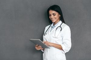 Arzt mit digitalem Tablet. Attraktive junge Ärztin im weißen Laborkittel, die an einem digitalen Tablet arbeitet und im Stehen vor grauem Hintergrund lächelt foto