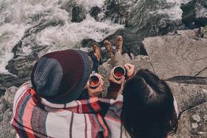 gemeinsam aufwärmen. Nahaufnahme von oben auf ein junges Paar, das mit einer Decke bedeckt ist, die Tassen hält, während es auf den Felsen in der Nähe des Flusses sitzt foto