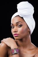 überzeugt von ihrer Schönheit. Schöne afrikanische Frau, die ein Kopftuch trägt und ihre Schulter berührt, während sie vor schwarzem Hintergrund steht foto