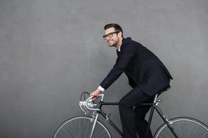 Ruhe bewahren und weiterfahren. Fröhlicher junger Geschäftsmann, der beim Radfahren vor grauem Hintergrund lächelt und wegschaut foto