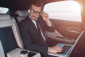 sein Geschäft planen. nachdenklicher junger Mann im vollen Anzug, der mit Laptop arbeitet und seine Brille anpasst, während er im Auto sitzt foto