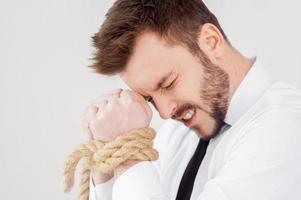 gefesselter Geschäftsmann. Seitenansicht eines frustrierten jungen Geschäftsmannes mit gefesselten Händen, die die Augen geschlossen halten, während sie vor grauem Hintergrund stehen foto