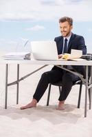 Arbeiten weit weg vom Büro. hübscher junger Mann in Abendkleidung, der am Laptop arbeitet und lächelt, während er am Tisch auf Sand sitzt foto