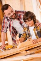 seinem Sohn alles über Zimmerei beizubringen. glücklicher junger männlicher Zimmermann, der seinen Sohn umarmt, während er in seiner Werkstatt mit Holz arbeitet foto