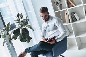 jung und klug. Der hübsche junge Mann ist eine elegante Freizeitkleidung, die etwas aufschreibt, während er im Büro arbeitet foto