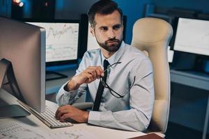 nachdenklicher junger Mann, der seine Brille hält und wegschaut, während er lange im Büro bleibt foto