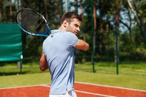 aufs Spiel konzentriert. hübscher junger mann im poloshirt, der tennisschläger hält und konzentriert aussieht, während er auf dem tennisplatz steht foto