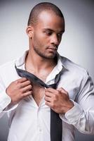 seine Krawatte ausziehen. Selbstbewusster junger schwarzer Mann, der seine Krawatte auszieht, während er vor grauem Hintergrund steht foto