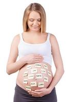 Namenswahl für ein Baby. Schöne schwangere Frau, die sich die Klebezettel mit Babynamen ansieht und lächelt, während sie isoliert auf Weiß steht foto
