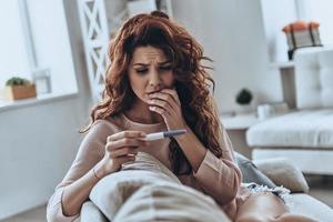 Was soll ich tun? Verängstigte junge Frauen sehen sich den Schwangerschaftstest an, während sie zu Hause auf dem Sofa sitzen foto