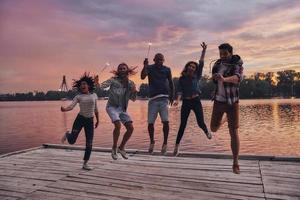 fühlt sich an wie Fliegen. in voller Länge von jungen Leuten in Freizeitkleidung, die lächeln und Funken halten, während sie auf den Pier springen foto