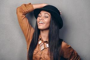 fröhliche Schönheit. Porträt einer schönen jungen, fröhlichen Afrikanerin mit Hut, die die Augen geschlossen hält und lächelt, während sie vor grauem Hintergrund steht foto