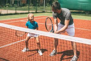Tennistraining. Fröhlicher Vater in Sportkleidung bringt seiner Tochter das Tennisspielen bei, während beide auf dem Tennisplatz stehen foto