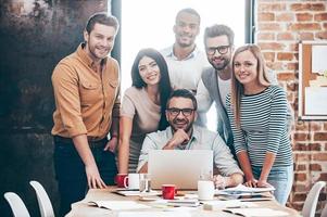 Perfektes kreatives Team. Gruppe von sechs fröhlichen jungen Leuten, die mit einem Lächeln in die Kamera schauen, während sie sich im Büro an den Tisch lehnen foto