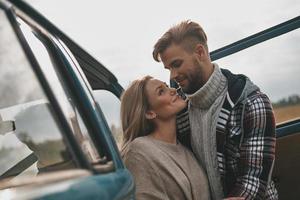 gerne zusammen reisen. Schönes junges Paar, das sich umarmt und lächelt, während es draußen in der Nähe des Minivans im Retro-Stil steht foto