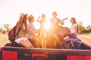 Sonnenuntergang treffen. Gruppe junger, fröhlicher Menschen, die ihren Roadtrip genießen, während sie zusammen im Pick-up-Truck sitzen foto