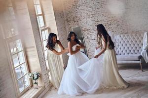 jedes Detail prüfen. Draufsicht in voller Länge von zwei attraktiven jungen Frauen, die ein Hochzeitskleid auf einer Braut justieren, während sie zusammen inear das Fenster stehen foto