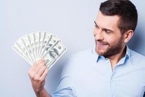 Geldliebhaber. glücklicher junger Mann, der die Papierwährung hält und sie betrachtet, während er vor grauem Hintergrund steht foto