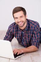 Surfen im Netz macht Spaß. hübscher junger Mann, der am Laptop arbeitet und lächelt, während er auf Hartholzboden liegt foto