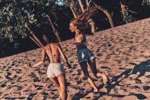tolle Zeit zusammen haben. Zwei attraktive junge Frauen in kurzen Hosen und Badebekleidung lächeln beim Laufen am Strand foto