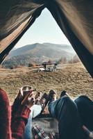 beste Reise aller Zeiten. Nahaufnahme eines jungen Paares, das morgens Kaffee trinkt, während es die Aussicht auf die Bergkette von seinem Zelt aus genießt foto