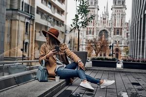 Attraktive junge Frau in Hut und Mantel, die über ihre Schulter schaut, während sie im Freien mit der Kirche im Hintergrund sitzt foto