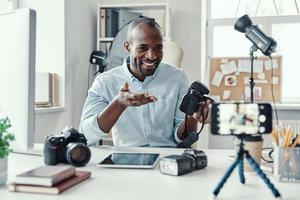 charmanter junger afrikanischer mann im hemd, der digitalkamera zeigt und etwas erzählt, während er social-media-videos macht foto