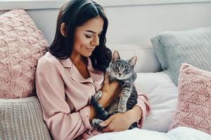 schöne junge frau im pyjama, die hauskatze lächelt und umarmt, während sie zu hause im bett ruht foto