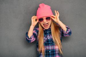 niedlich und emotional. Schönes kleines Mädchen, das ihre Sonnenbrille anpasst und den Mund offen hält, während es vor grauem Hintergrund steht foto