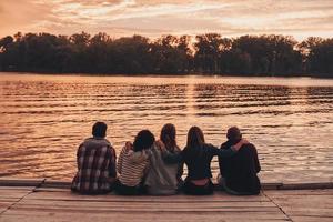 unter besten Freunden. Rückansicht junger Menschen in Freizeitkleidung, die sich umarmen, während sie auf dem Pier sitzen foto