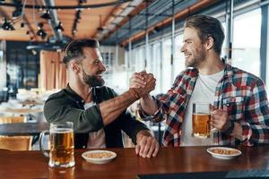 Fröhliche junge Männer in Freizeitkleidung, die sich die Hände schütteln und Bier trinken, während sie Zeit in der Kneipe verbringen foto