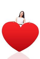 all meine Liebe gilt dir. Schöne junge asiatische Frau in hübschem Kleid, die sich an den riesigen roten, herzförmigen Valentinsgruß lehnt und lächelt, während sie vor weißem Hintergrund steht foto