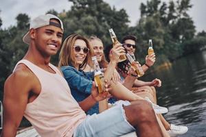 echte Freundschaft. Gruppe glücklicher junger Menschen in Freizeitkleidung, die lächeln und in die Kamera schauen, während sie auf dem Pier sitzen foto