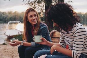 gutes Gespräch mit Freund. Zwei junge schöne Frauen in Freizeitkleidung lächeln und unterhalten sich beim Camping in der Nähe des Sees foto