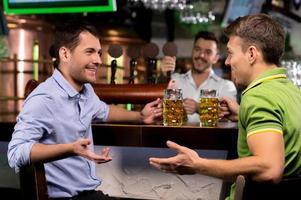 Zeit in der Bar verbringen. Zwei junge Männer trinken Bier und reden miteinander, während der Barkeeper im Hintergrund Bier klopft foto