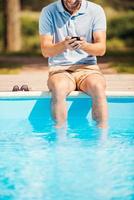 Mann am Pool entspannen. Zugeschnittenes Bild eines Mannes, der am Pool sitzt und etwas auf dem Handy tippt foto