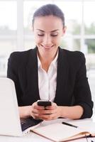 Geschäftsfrau mit Handy. Fröhliche junge Frau in Abendkleidung, die ein Handy hält und lächelt, während sie an ihrem Arbeitsplatz sitzt foto
