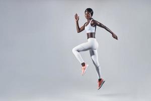 volle Länge der selbstbewussten jungen Afrikanerin in Sportkleidung, die vor grauem Hintergrund springt foto