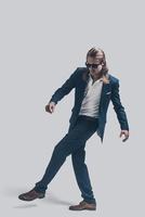 Eleganz in Bewegung. in voller Länge von einem hübschen jungen Mann in vollem Anzug und Sonnenbrille, der vor grauem Hintergrund posiert foto