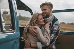 wahre Liebe ausdrücken. Schönes junges Paar, das sich umarmt und lächelt, während es draußen in der Nähe des Minivans im Retro-Stil steht foto