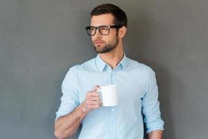 warten auf Inspiration. hübscher junger mann im hemd in den brillen, die tasse kaffee halten und weg schauen, während sie gegen grauen hintergrund stehen foto