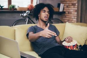 Knall junger afrikanischer Mann, der fernsieht und mit der Fingerpistole gestikuliert, während er zu Hause mit einem Eimer Popcorn auf der Couch sitzt foto