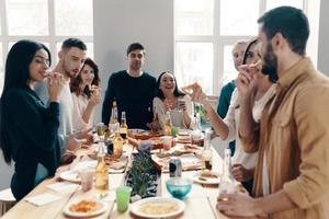 Zeit mit Freunden genießen. Gruppe junger Leute in Freizeitkleidung, die Pizza essen und lächeln, während sie drinnen eine Dinnerparty haben foto