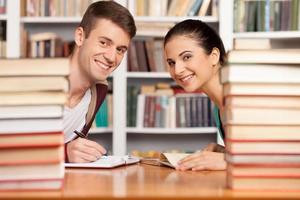 zusammen studieren. Fröhlicher junger Mann und Frau, die am Bibliothekstisch sitzen und aus den Bücherstapeln schauen foto